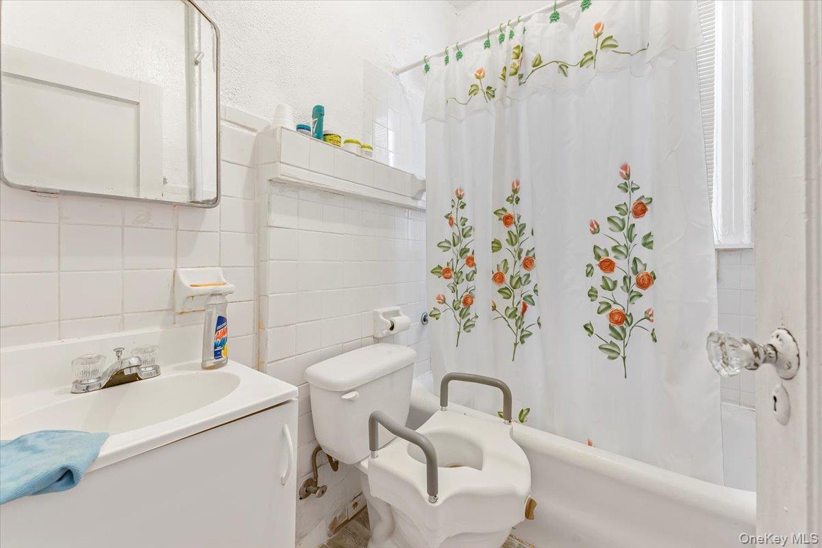 Bathroom with toilet, shower / bathtub combination with curtain, tile walls, and vanity