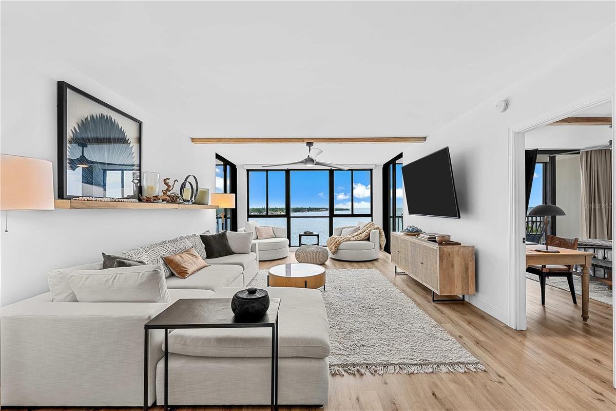 Open-concept living room with sectional seating, swivel lounge chairs, custom wood shelf, and panoramic Tampa Bay views through walls of glass.