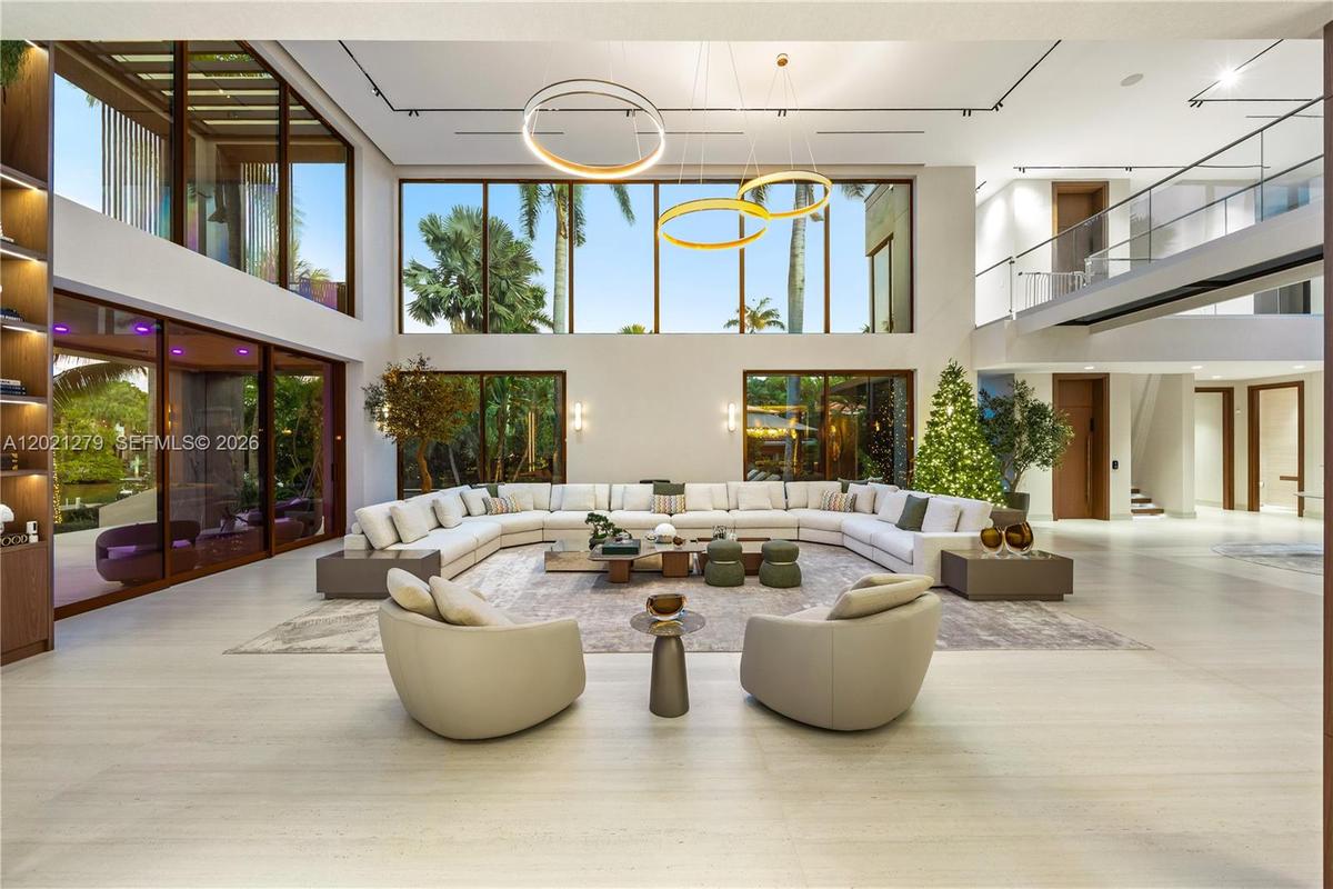 Grand double-height living room with wraparound sectional seating, light stone floors, dual ring chandelier, full-wall glass overlooking tropical wate