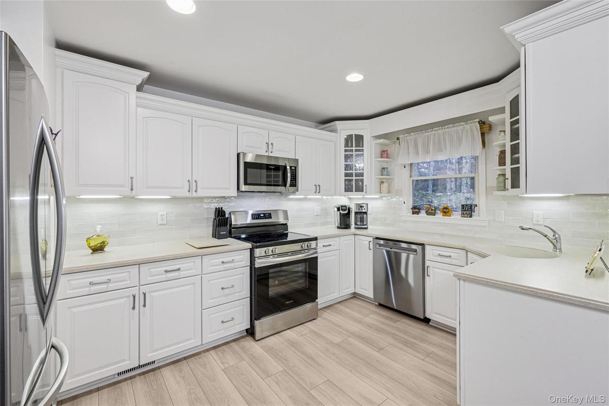 Kitchen featuring appliances with stainless steel finishes, white cabinets, glass insert cabinets, decorative backsplash, and light stone countertops