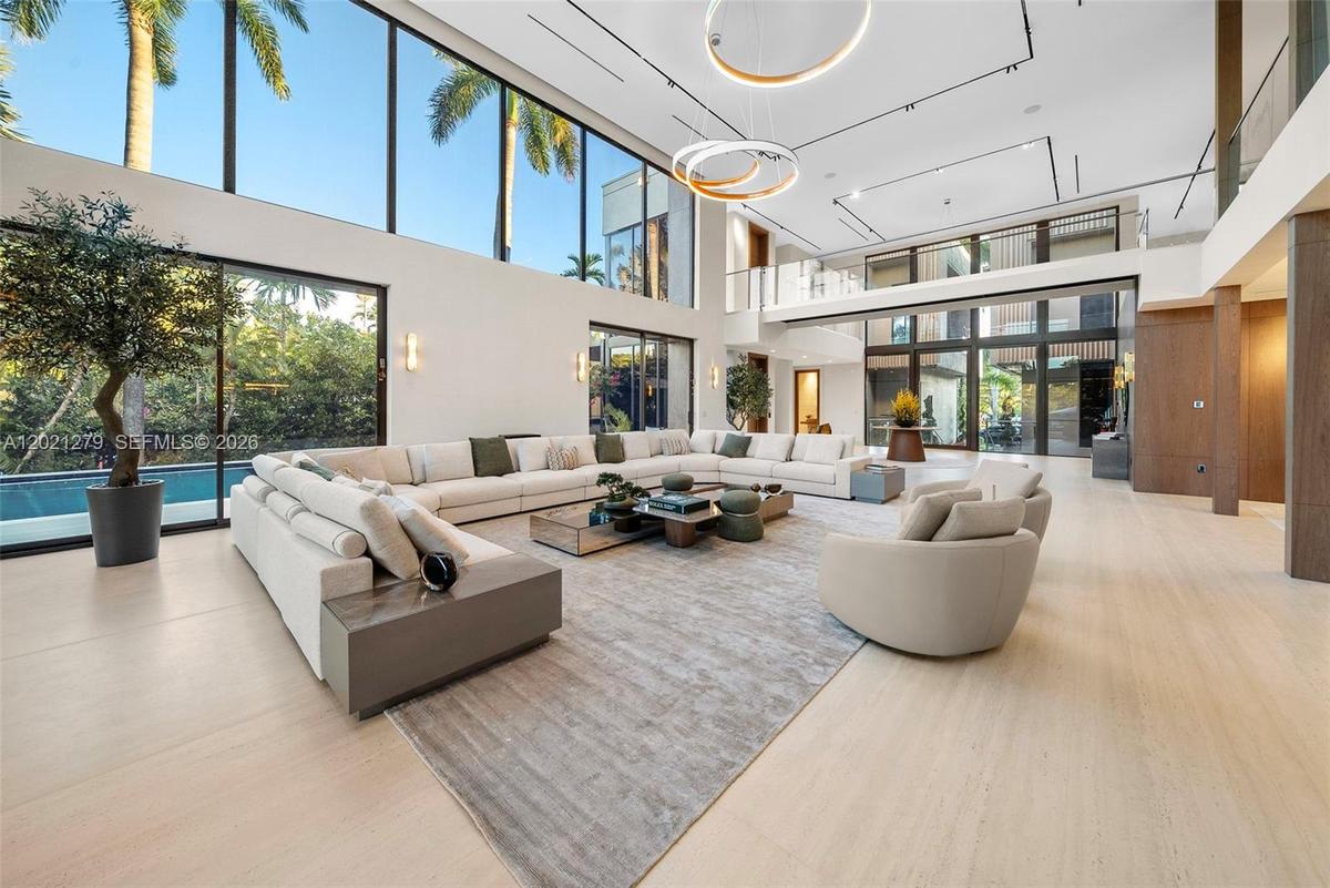 Expansive double-height living room with oversized curved sectional, ring chandelier, floor-to-ceiling glass with direct pool and tropical garden view