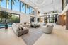 Expansive double-height living room with oversized curved sectional, ring chandelier, floor-to-ceiling glass with direct pool and tropical garden view