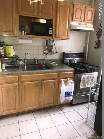Kitchen featuring stainless steel gas range oven, light tile patterned floors, dark countertops, and brown cabinets