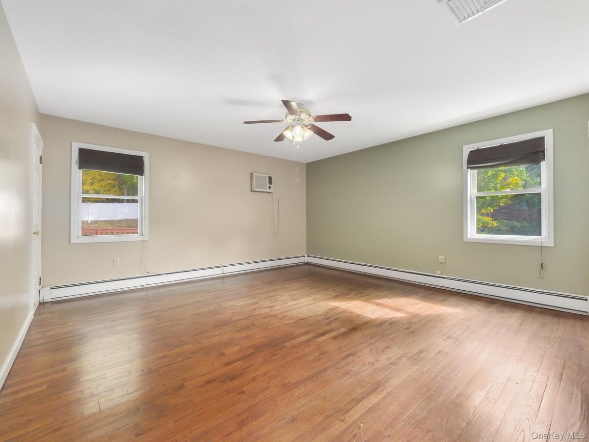 Hardwood flooring, ceiling fan, window-mounted air conditioning unit, and multiple windows with roller shades