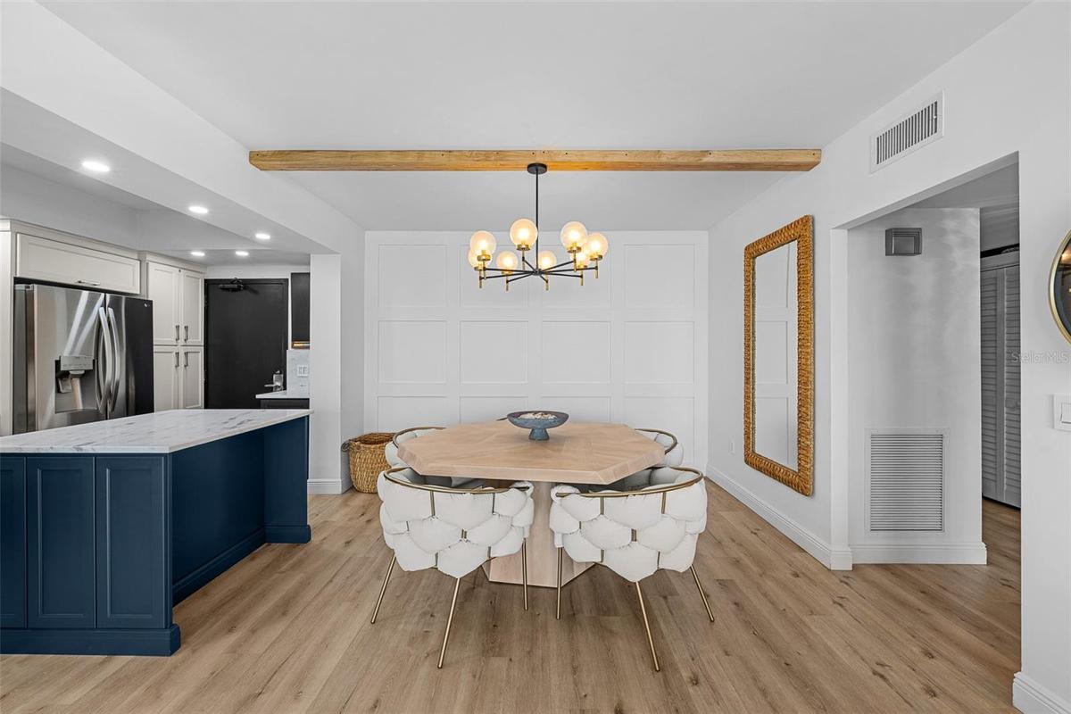 Dining area with a custom octagonal hardwood table, brass-and-glass chandelier, navy kitchen island in the background, and a panel-board accent wall.